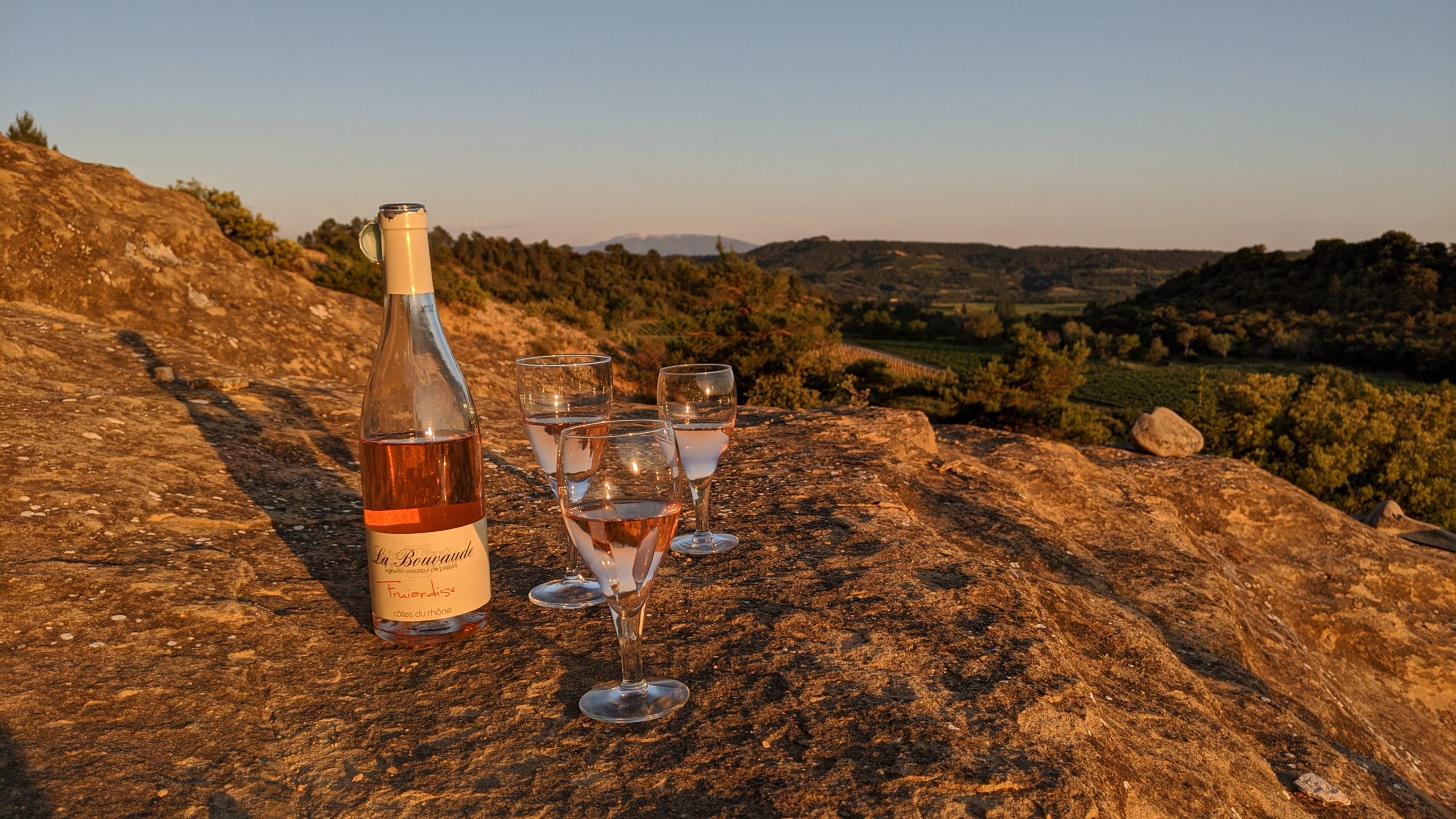 Une bouteille de rosé et trois verres de vin posés sur un rocher au coucher de soleil
